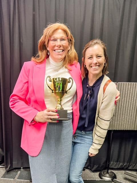 Two women smiling; Deanna Jones in a pink blazer holding a gold trophy, the other in a beige cardigan standing close beside her, with a black backdrop behind them.