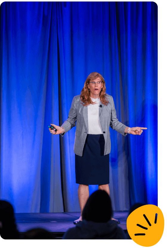 Deanna Jones, in a blazer and skirt, stands on stage speaking and gesturing with both hands, framed by a blue curtain backdrop and an attentive audience in the foreground.