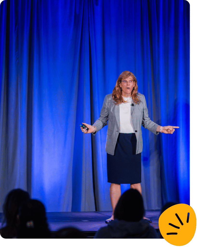 Deanna Jones, dressed in business attire, stands on stage speaking, with a blue curtain backdrop and an audience in the foreground.