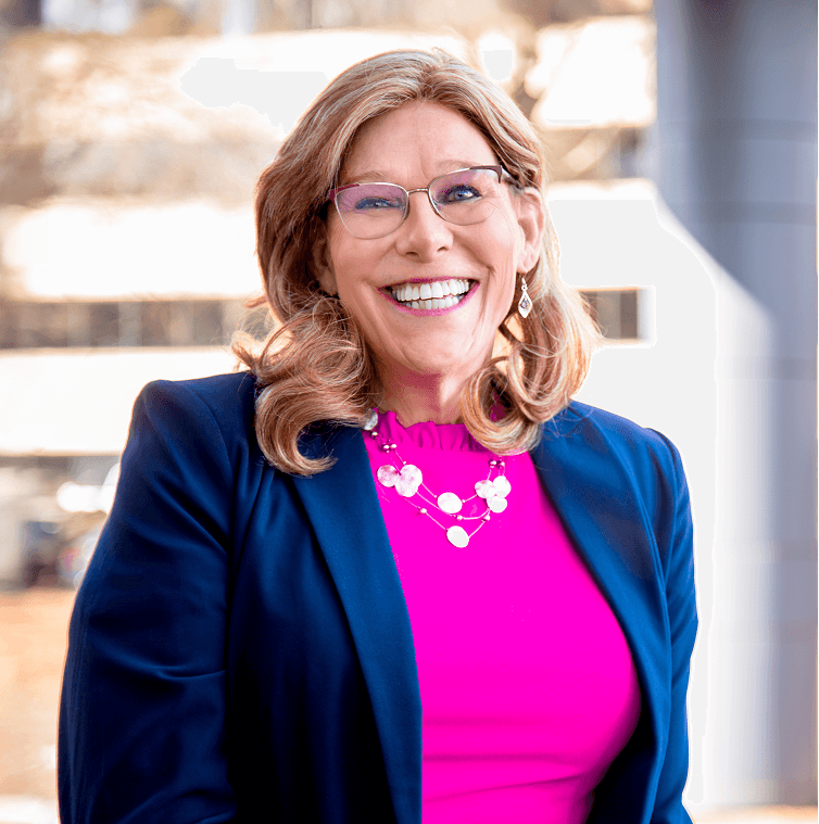 Smiling woman, Deanna Jones, with shoulder-length blonde hair, wearing glasses, a blue blazer, a bright pink top, and a pink beaded necklace, stands indoors against a blurred background.