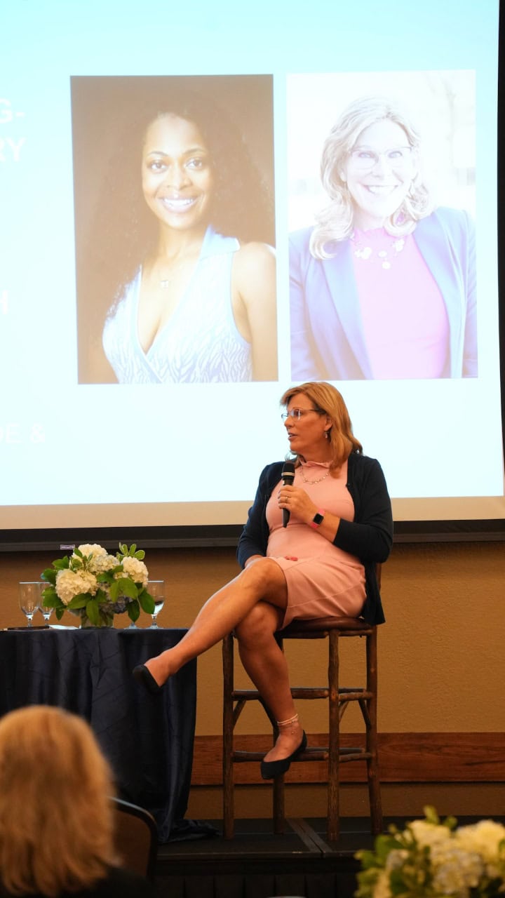 Deanna Jones sits on a high chair holding a microphone, with projected portraits of two women displayed on a screen behind her.