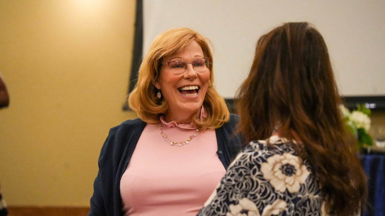 Deanna Jones, a woman with blonde hair and glasses, smiles and chats with another woman with long brown hair in a patterned top at an indoor event.