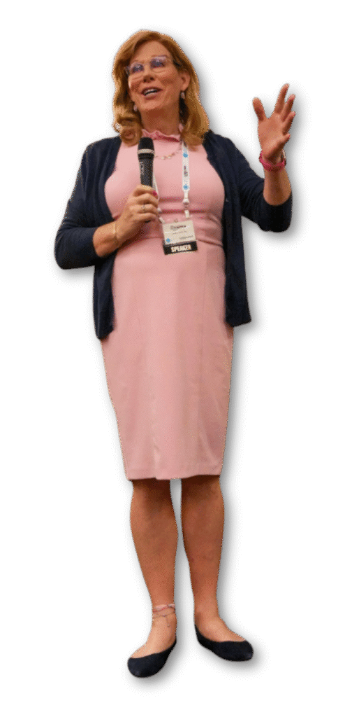 A woman in a pink dress and navy cardigan, identified as Deanna Jones by her "Speaker" name badge, holds a microphone and gestures while addressing the event.