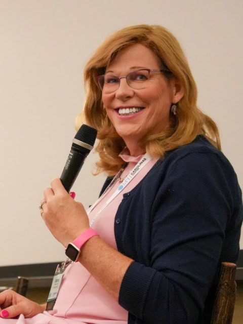 Deanna Jones, with shoulder-length hair, wearing glasses and a cardigan, smiles while holding a microphone and sitting indoors.